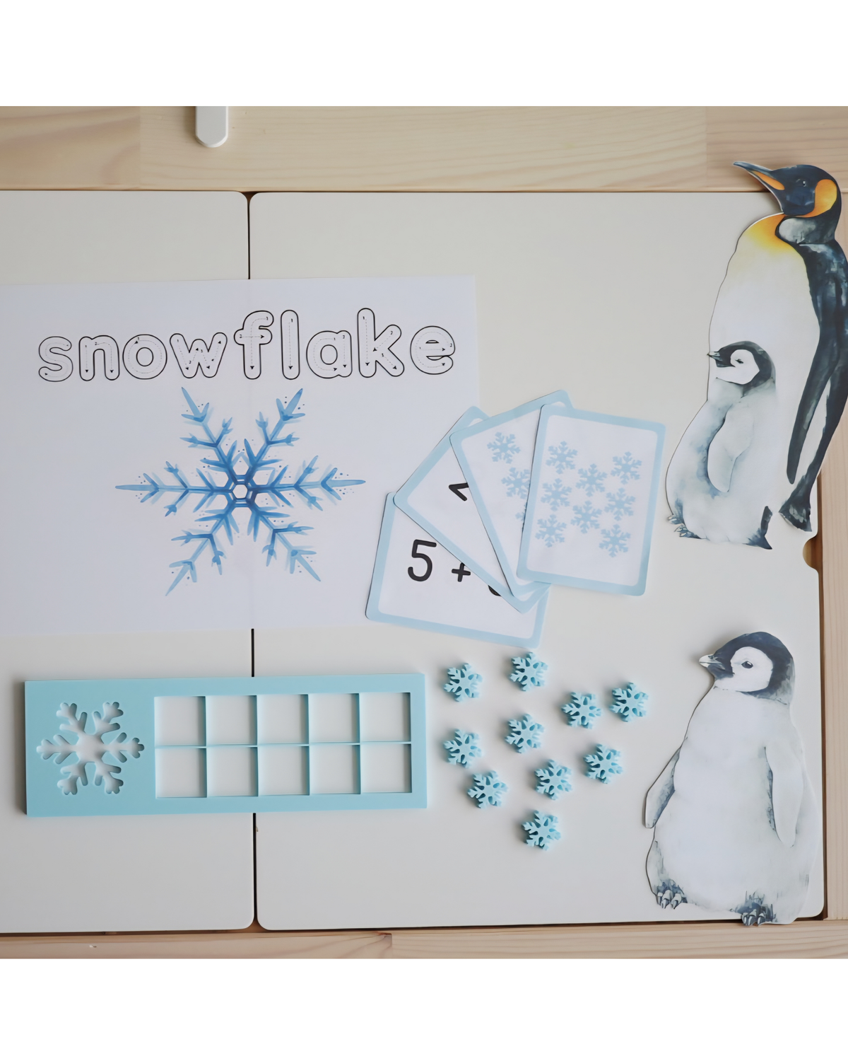 Tabliczka edukacyjna Snowflake 10 Frame z niebieskimi liczmanami w kształcie śnieżynek, otoczona zimowymi kartami i grafikami pingwinów – zestaw do nauki liczenia w stylu Montessori.