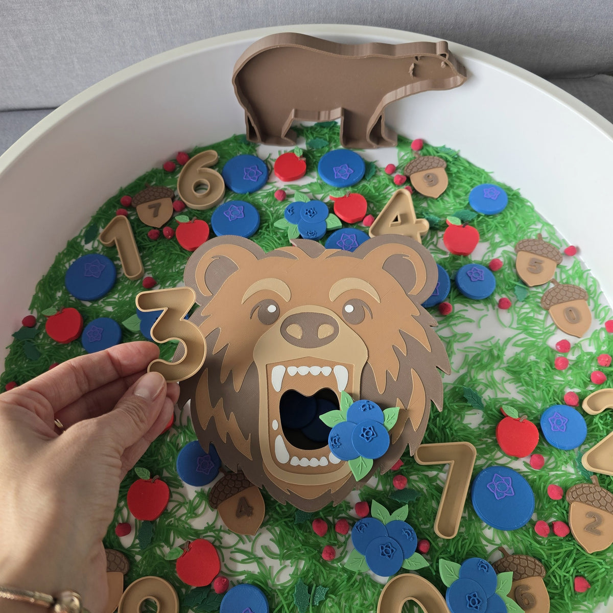 Child feeds a 3D printed bear a blueberry, numbered acorns and apples in a forest sensory bin