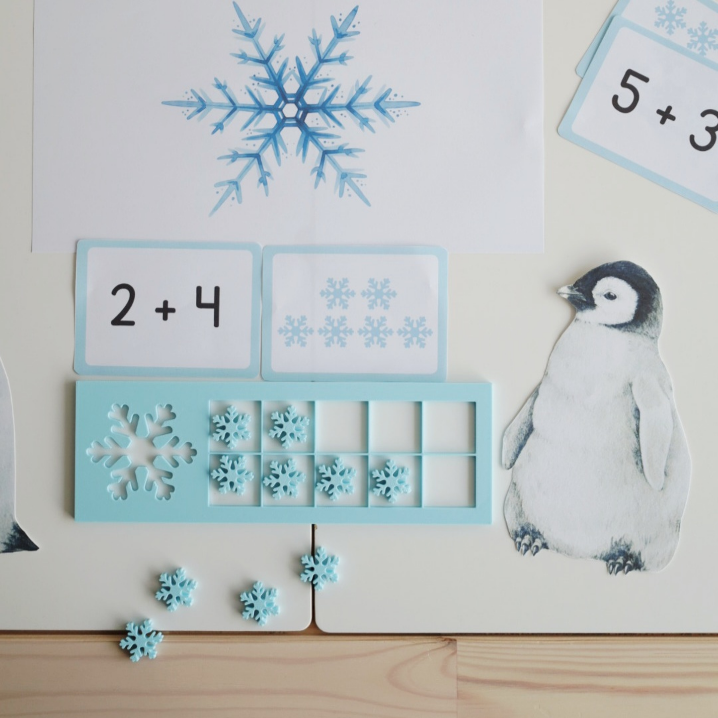 Zimowa scenka edukacyjna z tabliczką Snowflake 10 Frame, kartami do liczenia i grafiką małego pingwina – zachęta do sensorycznej nauki liczenia.