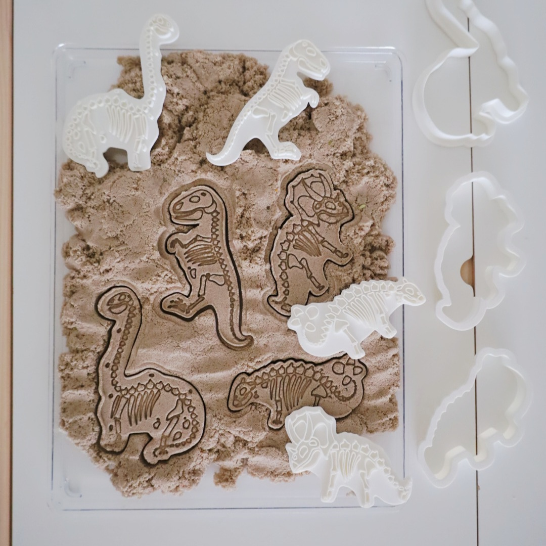 Dinosaur-themed sensory stamps and cutters creating fossil-style impressions in kinetic sand, including T-rex, brontosaurus, triceratops and stegosaurus shapes.