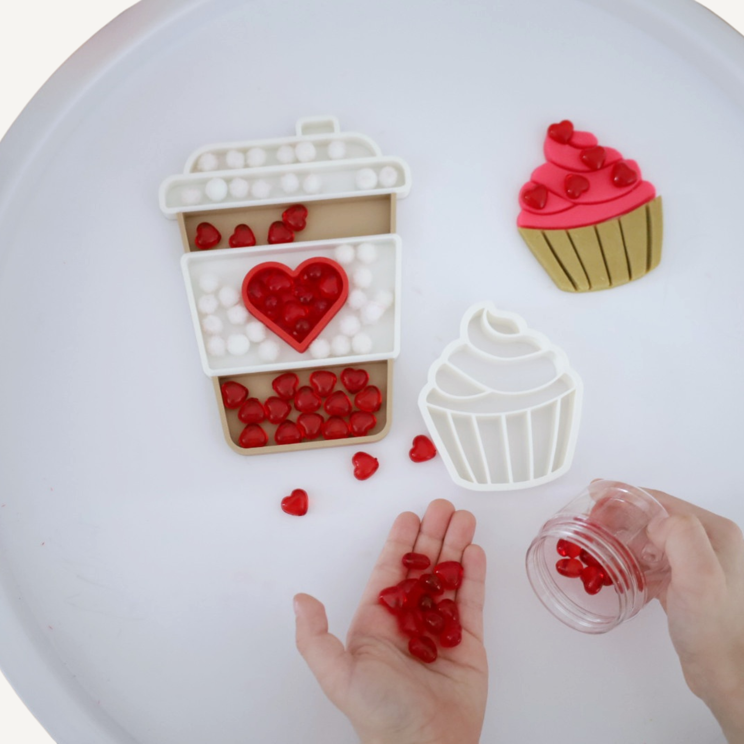 Child’s hands play with red heart-shaped beads, filling Montessori-style Valentine's themed trays shaped like a coffee cup and cupcakes.