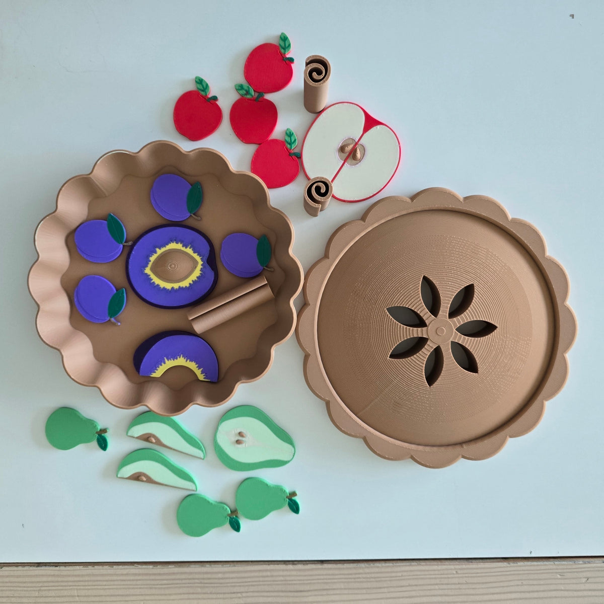 Montessori autumn fruit pie play set with pie crust lid, 3D printed apples, pears, plums, fruit halves and cinnamon sticks laid out on a white table