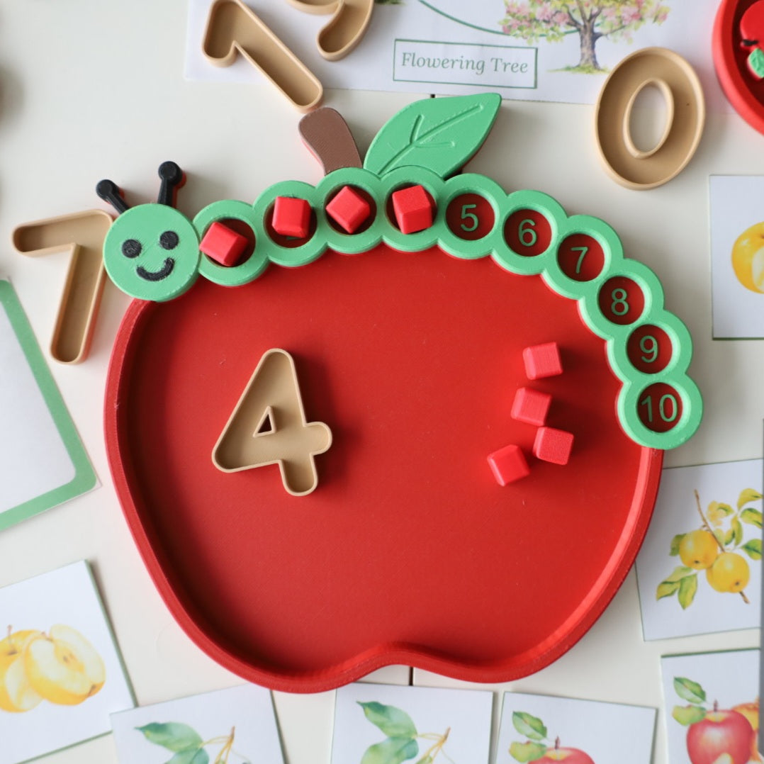 Red 3D printed apple counting tray with a smiling green caterpillar counting track numbered 1 to 10, red counting cubes placed in the first four circles, and a wooden-look number 4 inside the tray next to four loose red cubes, surrounded by apple picture cards.