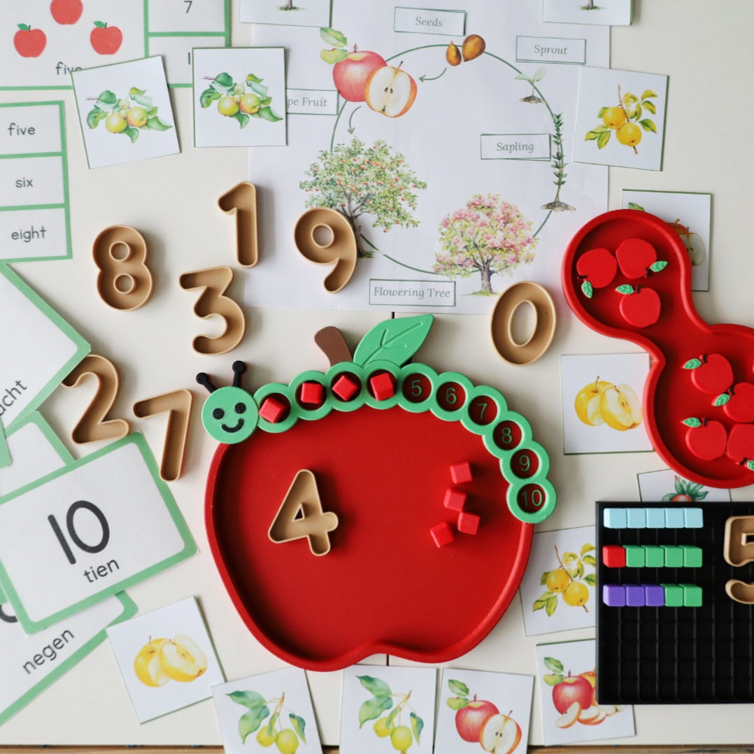 Montessori apple-themed math activity with a red apple counting tray and green caterpillar number track, mini numbers 0 to 9, red counting cubes, a red part-part-whole tray with apple counters, and an apple tree life cycle poster with number cards in English and Dutch.